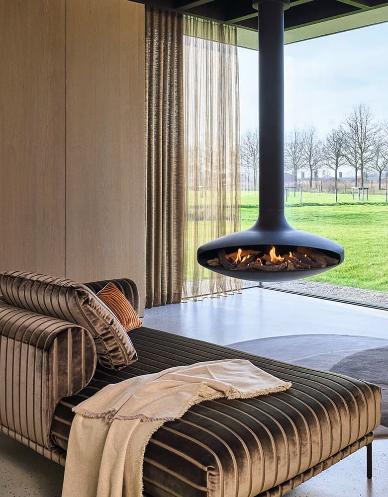Eine Aufnahme von einem Wohnzimmer mit einer bronzefarbenen Couch und einem schwebenden Kamin vor dem bodentiefen Fenster.