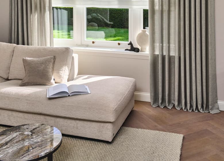Eine Aufnahme von einer Wohnzimmersituation mit einem beigen Sofa und einem gräulichen Vorhang vor dem Fenster.