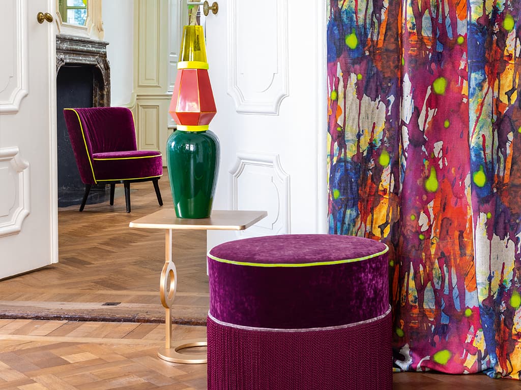 Das Bild zeigt einen lila Pouf vor einem multicoloren Vorhang. Der Raum hat eine hohe Wandhöhe, erkennbar an den Doppeltüren, die ins Kaminzimmer führen. Die auffälligen Farben des Vorhangs harmonieren hervorragend mit der eleganten Einrichtung.  