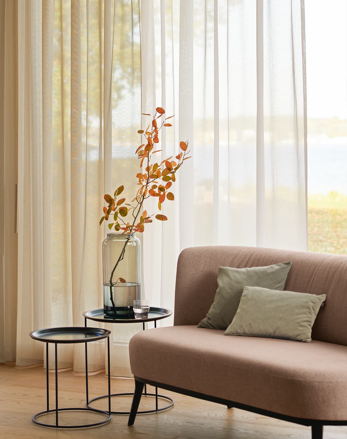 Das Bild zeigt ein Sofa mit Kissen vor einer transparenten Gardine. Im Raum steht ein kleines braunes Sofa sowie zwei unterschiedlich hohe Beistelltische. Die Atmosphäre ist warm, unterstützt durch die gelb-braunen Gardinen.