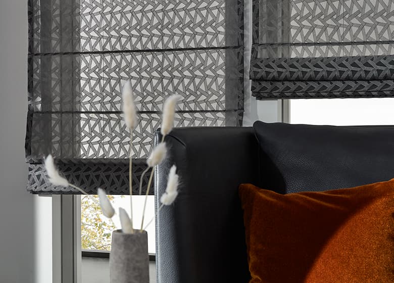 Dunkles Ledersofa mit einem orangenen Samtkissen und grauen transparenten Faltrollos im Hintergrund mit 3D-Effekt und Stickereien