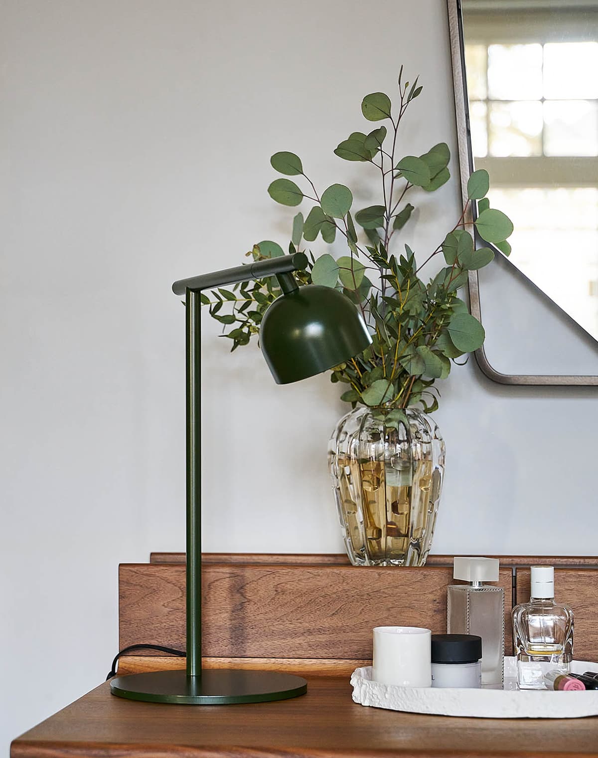 Tischleuchte in grün auf einem Schreibtisch neben Kosmetikprodukten. Hinter der Lampe steht eine Vase mit Wasser und Pflanze.