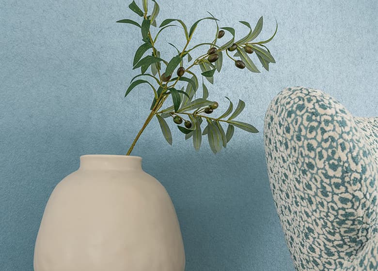 Das Bild zeigt eine Nahaufnahme einer blauen Tapete. Vor der Wand steht eine Vase mit einem Dekozweig. Rechts im Bild ist die Lehne eines blau-weißen Sessels zu erkennen.