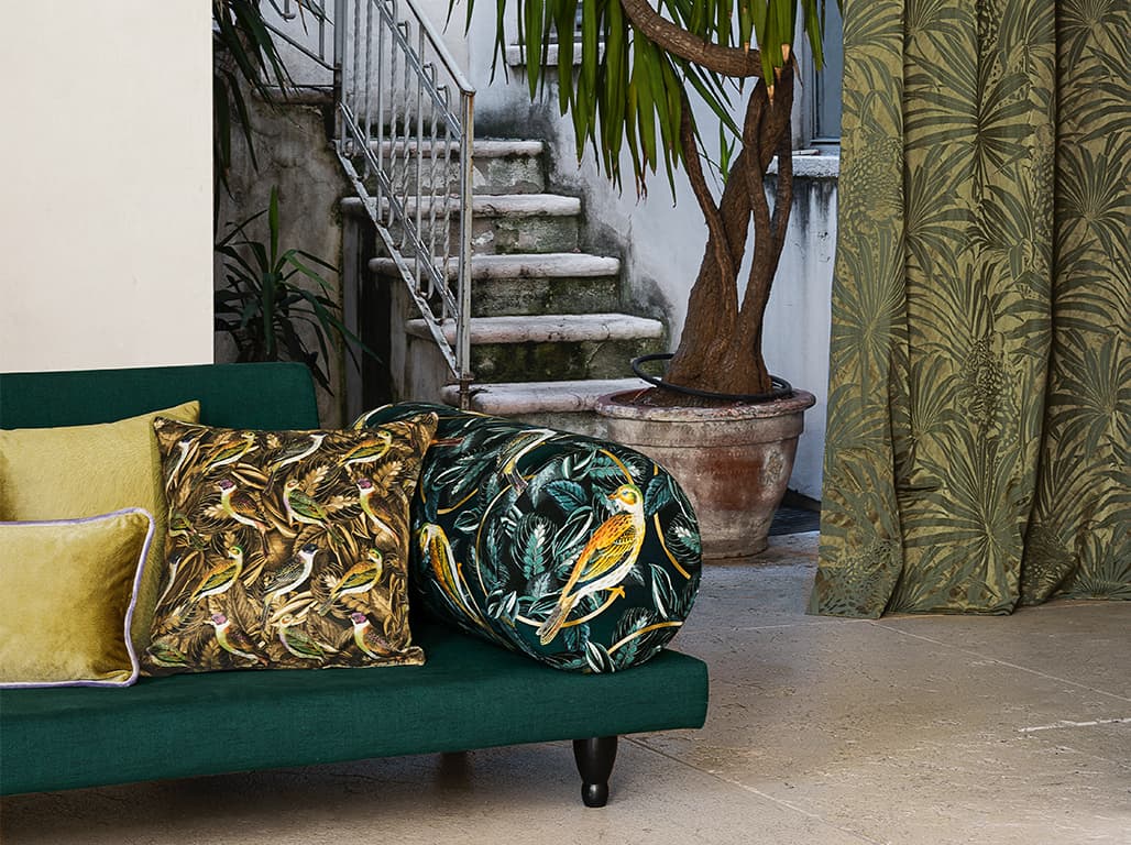 Das Bild zeigt ein grünes Sofa mit floralen und gelben Kissen. Rechts hängt ein grüner Vorhang, dahinter ist ein rustikaler Treppenaufstieg. Die Szene wirkt einladend und gemütlich.
