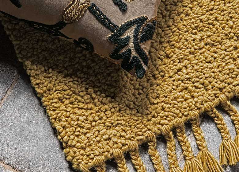 Das Bild zeigt einen gelben Teppich mit klarer Struktur und einem Kissen darauf. Der Teppich auf dem Steinboden schafft einen warmen Kontrast und vermittelt Gemütlichkeit.