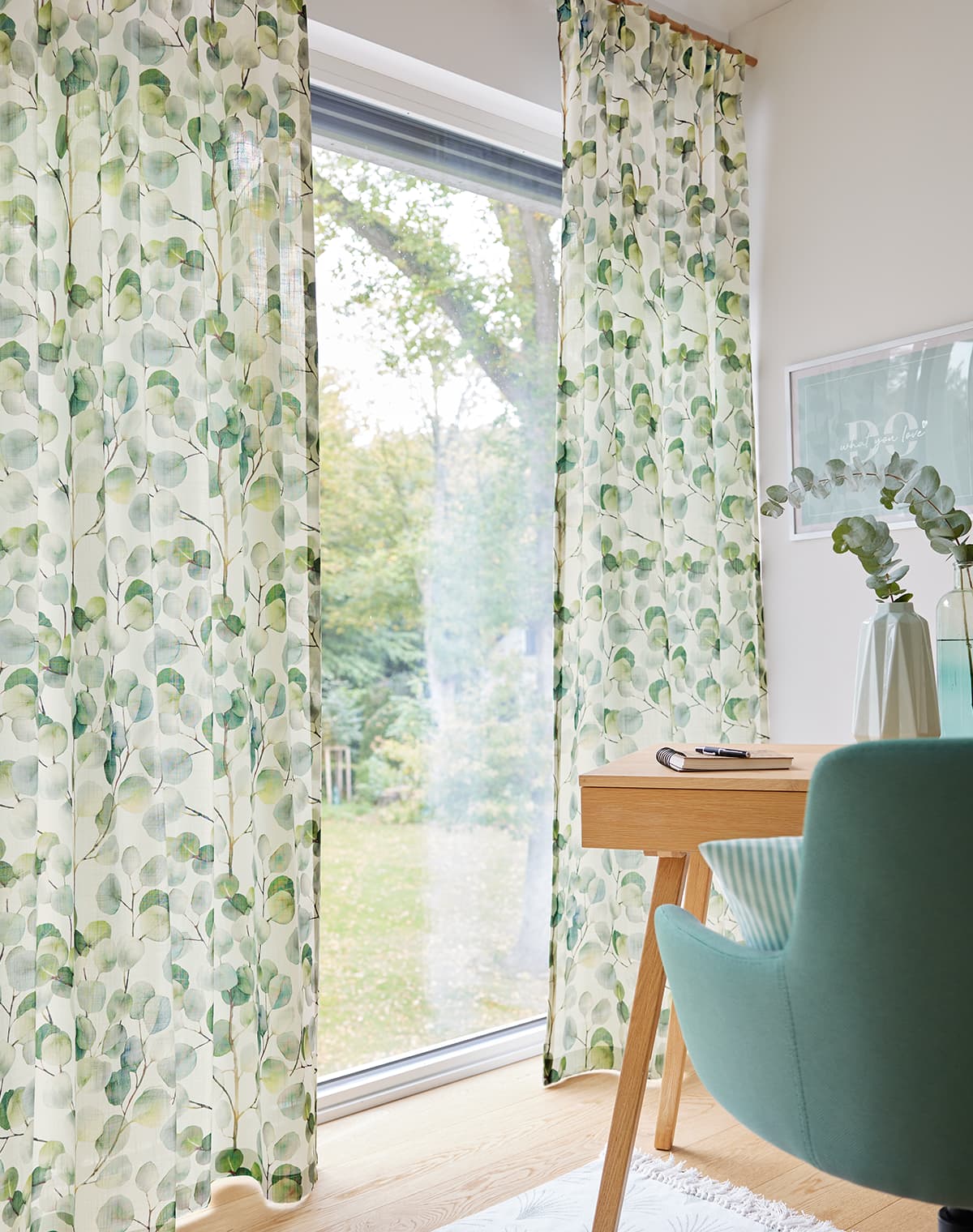 Das Bild zeigt eine Gardine mit grünem floralem Design, die einen hellen Arbeitsplatz am Fenster umrahmt. Die Farbgestaltung ist überwiegend in hellen Grüntönen gehalten, was eine frische und einladende Atmosphäre schafft.