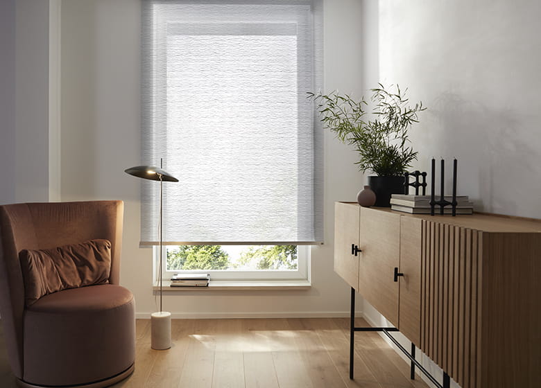 Lounge mit braunem Sessel, Sideboard aus Holz und einem hellgrauen Rollo mit Wellenmuster am Fenster