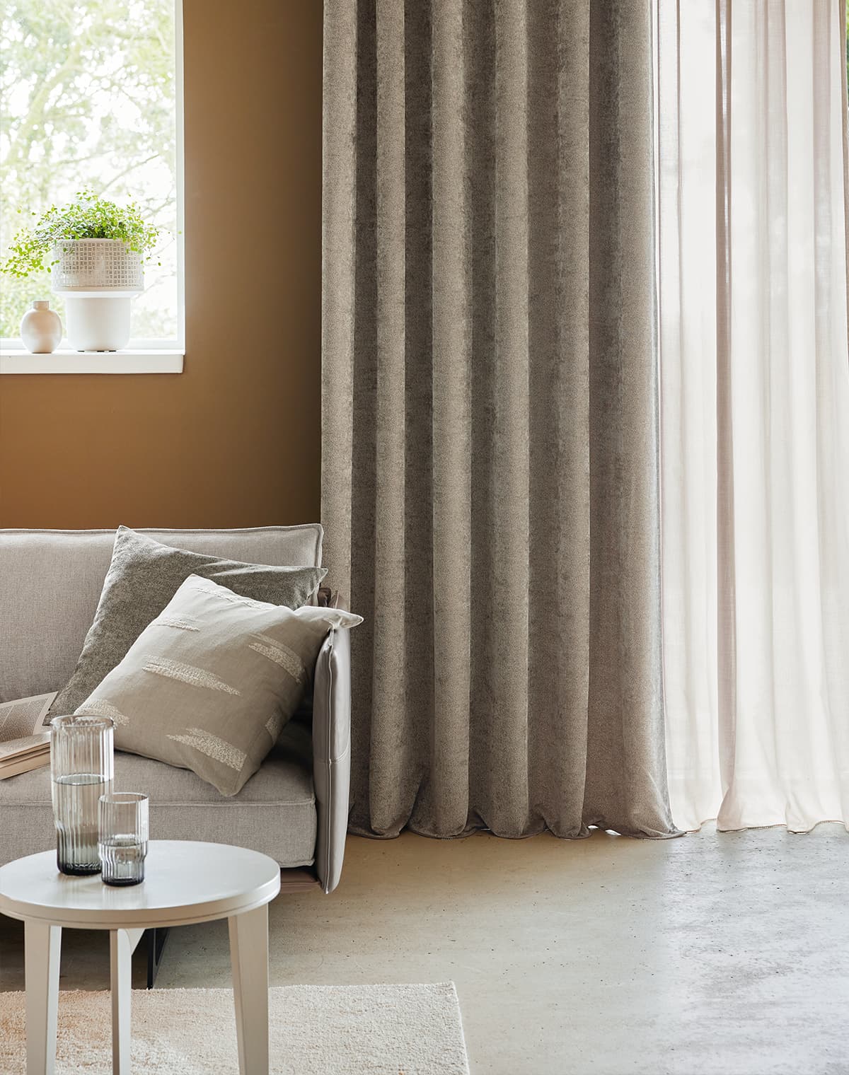 Das Zimmer hat ein hellbraunes Sofa mit gleichfarbigen Kissen, einen Beistelltisch und einen weißen Teppich. Ein raumhoher Vorhang schafft Gemütlichkeit.