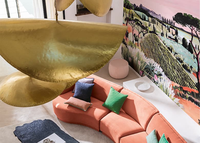 Das Bild zeigt ein koralfarbenes Sofa vor einer bunten Tapete, die dem Raum Lebendigkeit verleiht. Die unterschiedlich farbigen Kissen auf dem Sofa spiegeln die Farben der Tapete wieder.