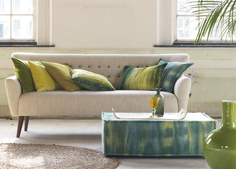 Das Bild zeigt einen hellen Raum mit einem beigen Sofa, das mit bunten Kissen dekoriert ist. Ein Teppich, ein Beistelltisch und verschiedene Accessoires tragen zu einer angenehmen Stimmung bei.