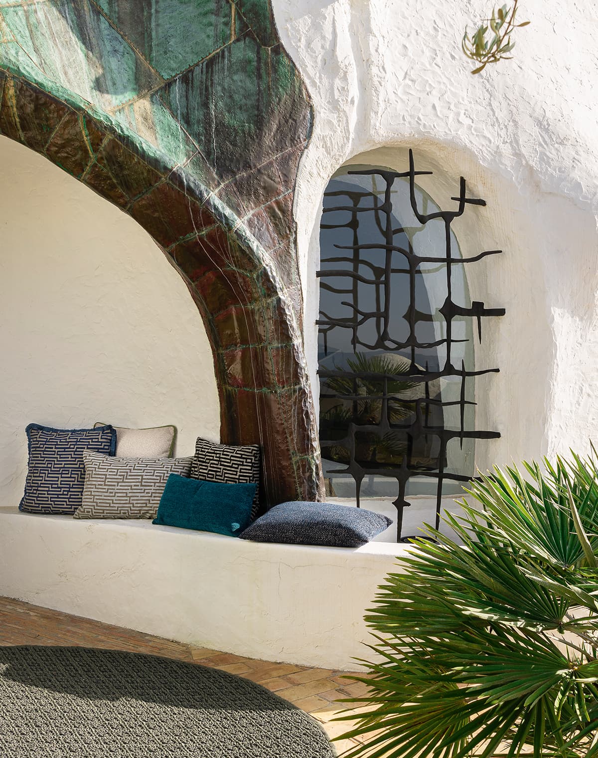 Das Bild zeigt eine in die Architektur integrierte Bank mit bunten, gemusterten Kissen. Ein runder Teppich liegt auf dem Boden, und eine Pflanze bringt frisches Grün ins Bild. Die geschwungene Architektur harmoniert gut mit den Kissen und dem Teppich.