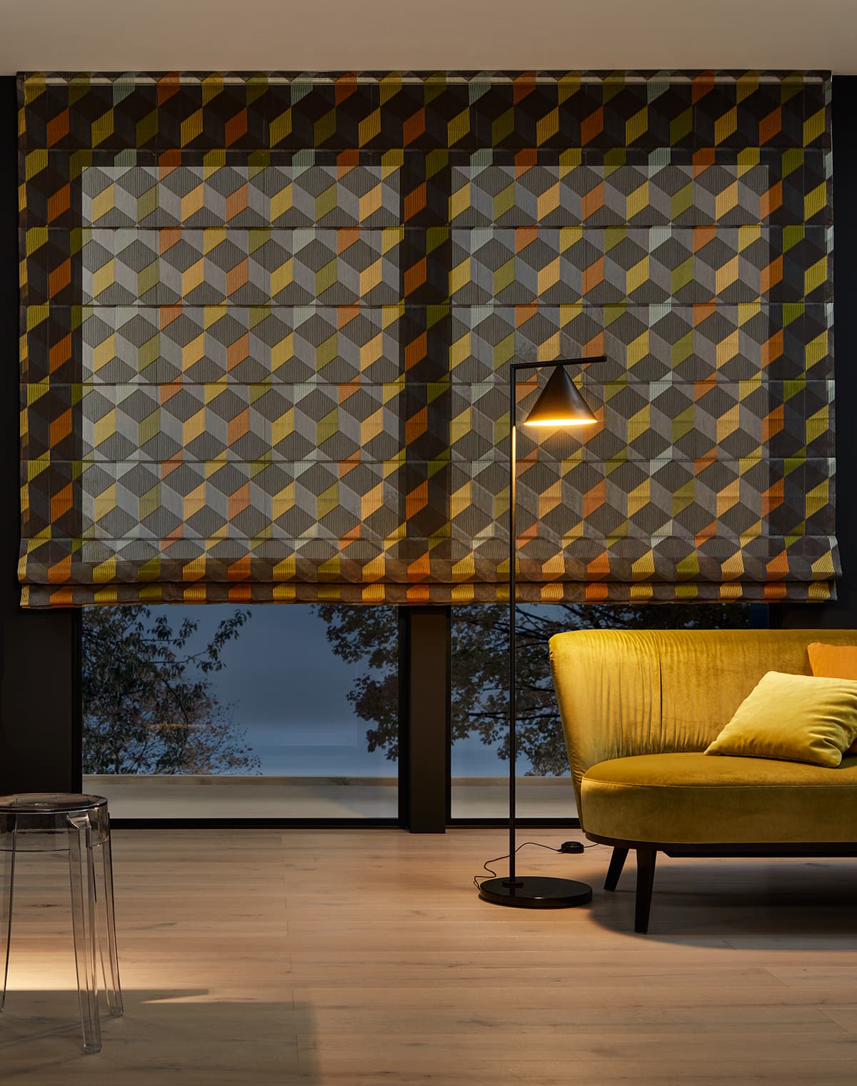 Transparentes Faltrollo in dreifarbigem Rautemuster an einem bodentiefen Fenster und gelbem Sofa.