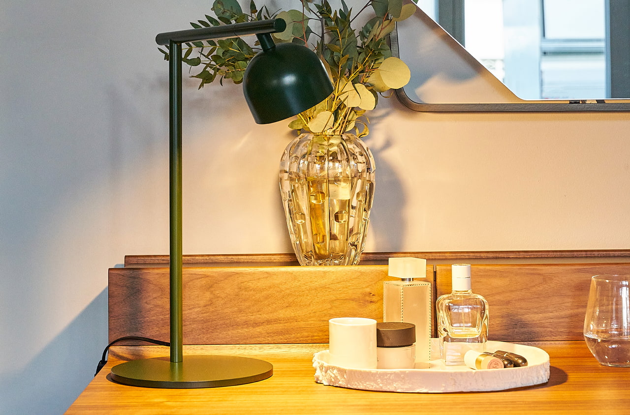 Tischleuchte in grün auf einem Schreibtisch neben Kosmetikprodukten. Hinter der Lampe steht ein Blumentopf mit Wasser und Pflanze.