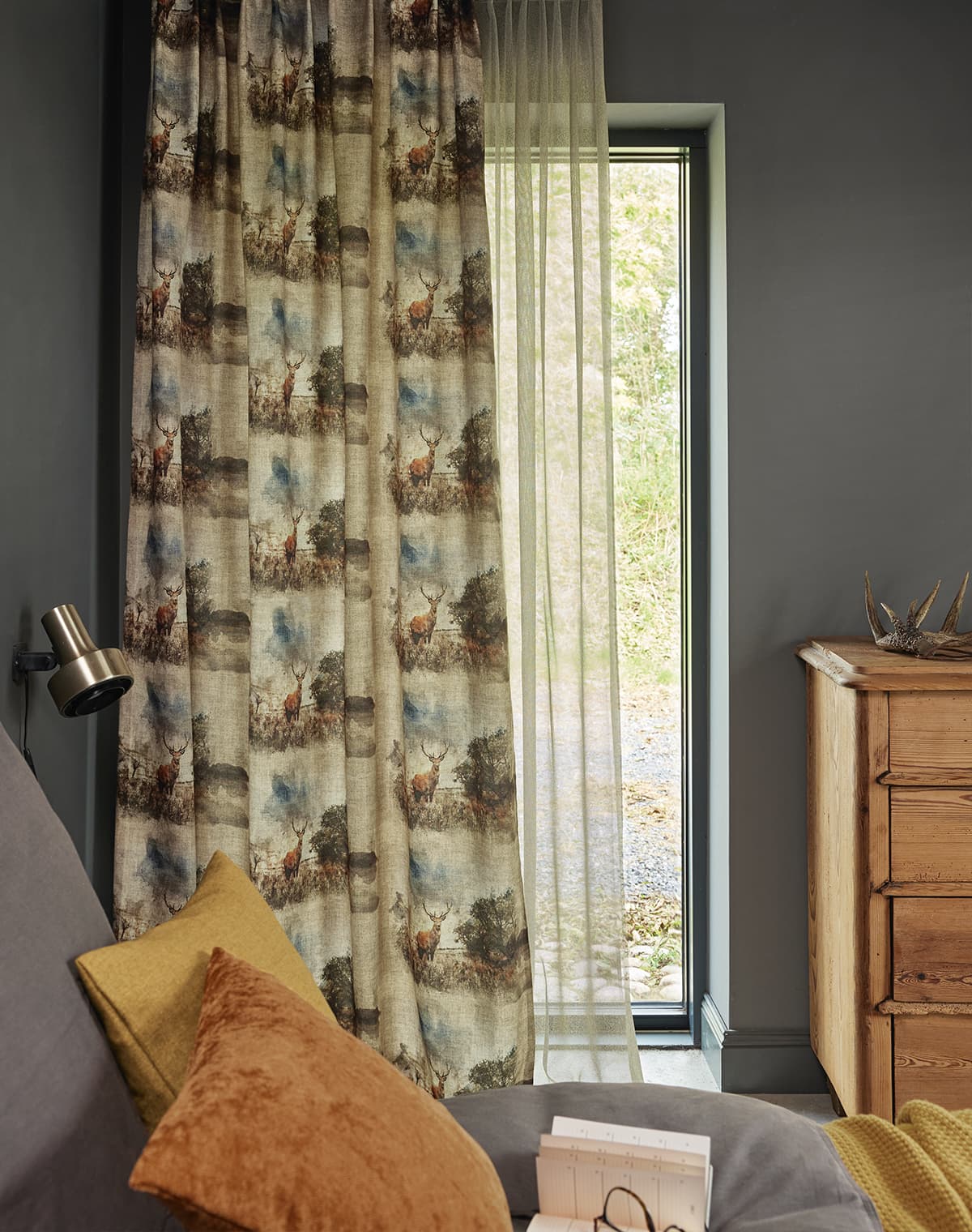 Das Bild zeigt eine Gardine mit Waldmotiven, die am Fenster hängt. Im Ausschnitt des Bildes ist ein Bett zu sehen, das mit einem orangefarbenen und einem gelben Kissen dekoriert ist, was dem Raum eine angenehme Atmosphäre verleiht.