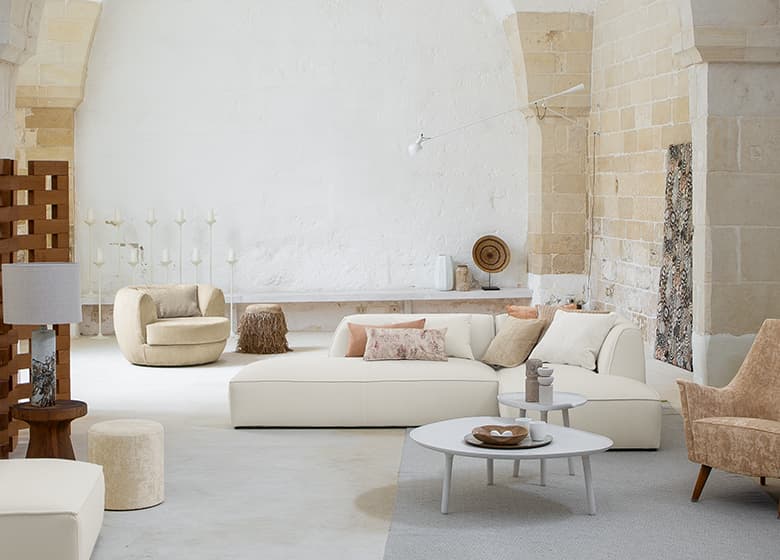 Das Bild zeigt einen großzügigen Raum mit vielfältiger Wohnzimmer-Einrichtung. Ein Sofa und Sessel in Weiß, Beige und Braun sind prominent platziert. Zahlreiche Accessoires im Raum tragen zur farblichen Gestaltung und gemütlichen Atmosphäre bei.