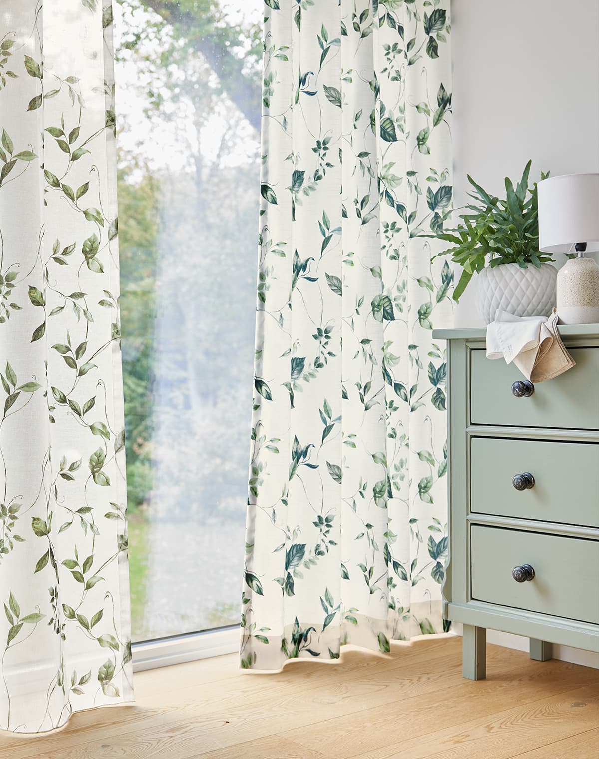 Das Bild zeigt eine Gardine mit hellgrünem und eine mit dunkelgrünem floralem Muster. Die hellere grüne Kommode rechts im Bild unterstützt das florale Design, während der warme Holzboden eine gemütliche Atmosphäre schafft.