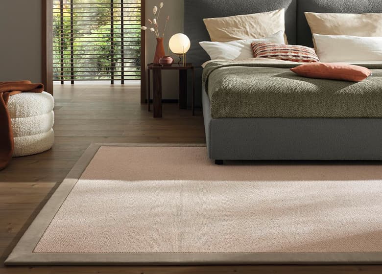 Eine Schlafzimmersituation mit einem dunkelgrünen Bett. Unter dem Bett liegt ein beiger Teppich mit dunklerem Rand.