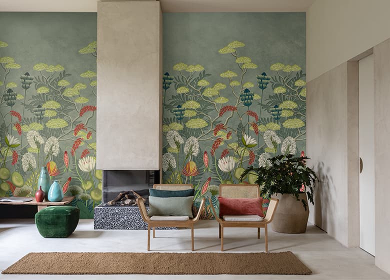 Das Bild zeigt eine Tapete mit Blumenprint in einem wohnlich eingerichteten Raum mit Kamin, Stuhl und Hocker. Die grüne Wand harmoniert gut mit dem weißen Boden und schafft eine einladende Atmosphäre.