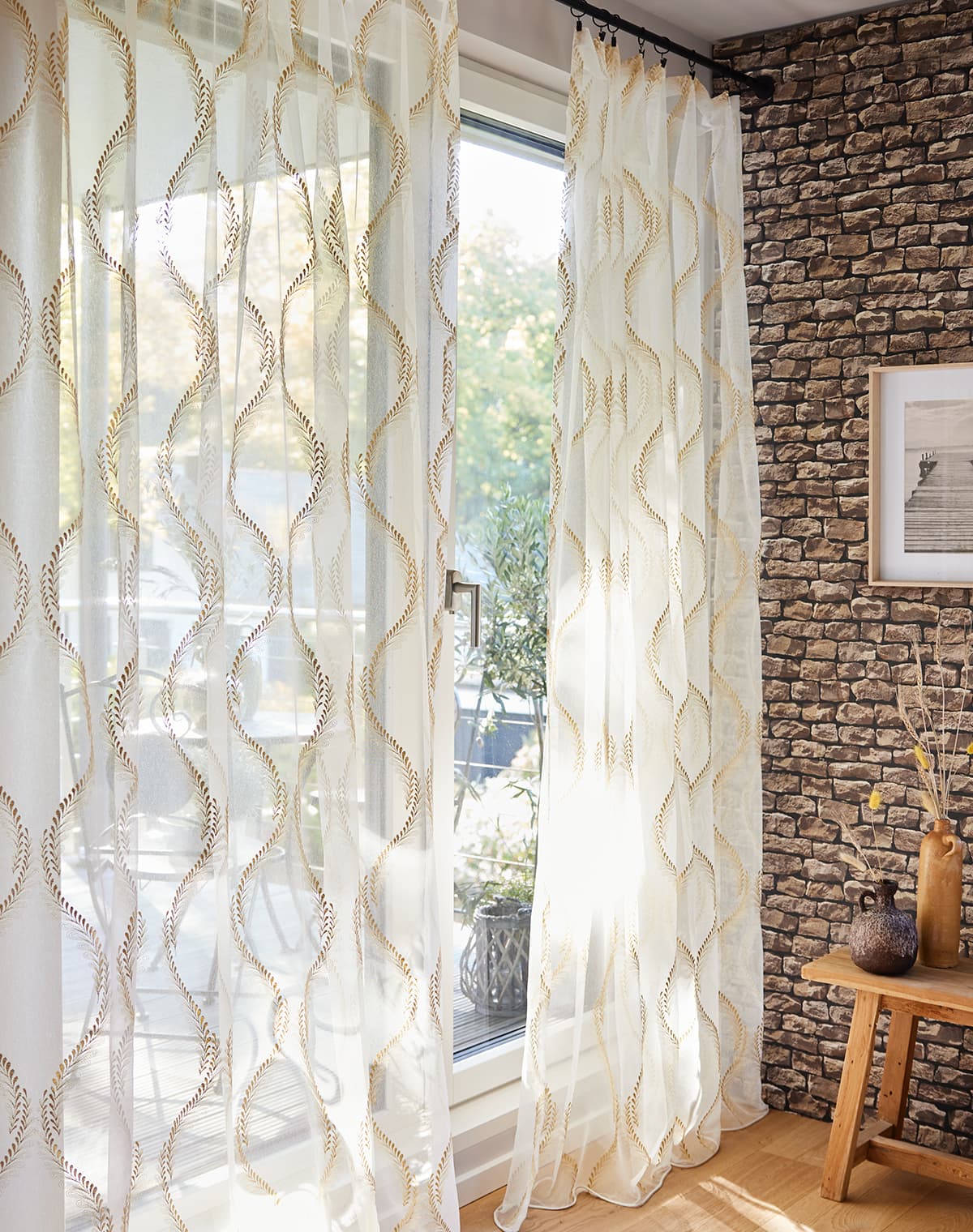 Das Bild zeigt eine weiße Gardine mit braunem floralem Streifendesign. Die Kombination mit der rustikalen Wand und den Beistellelementen vermittelt ein ländliches Gefühl.