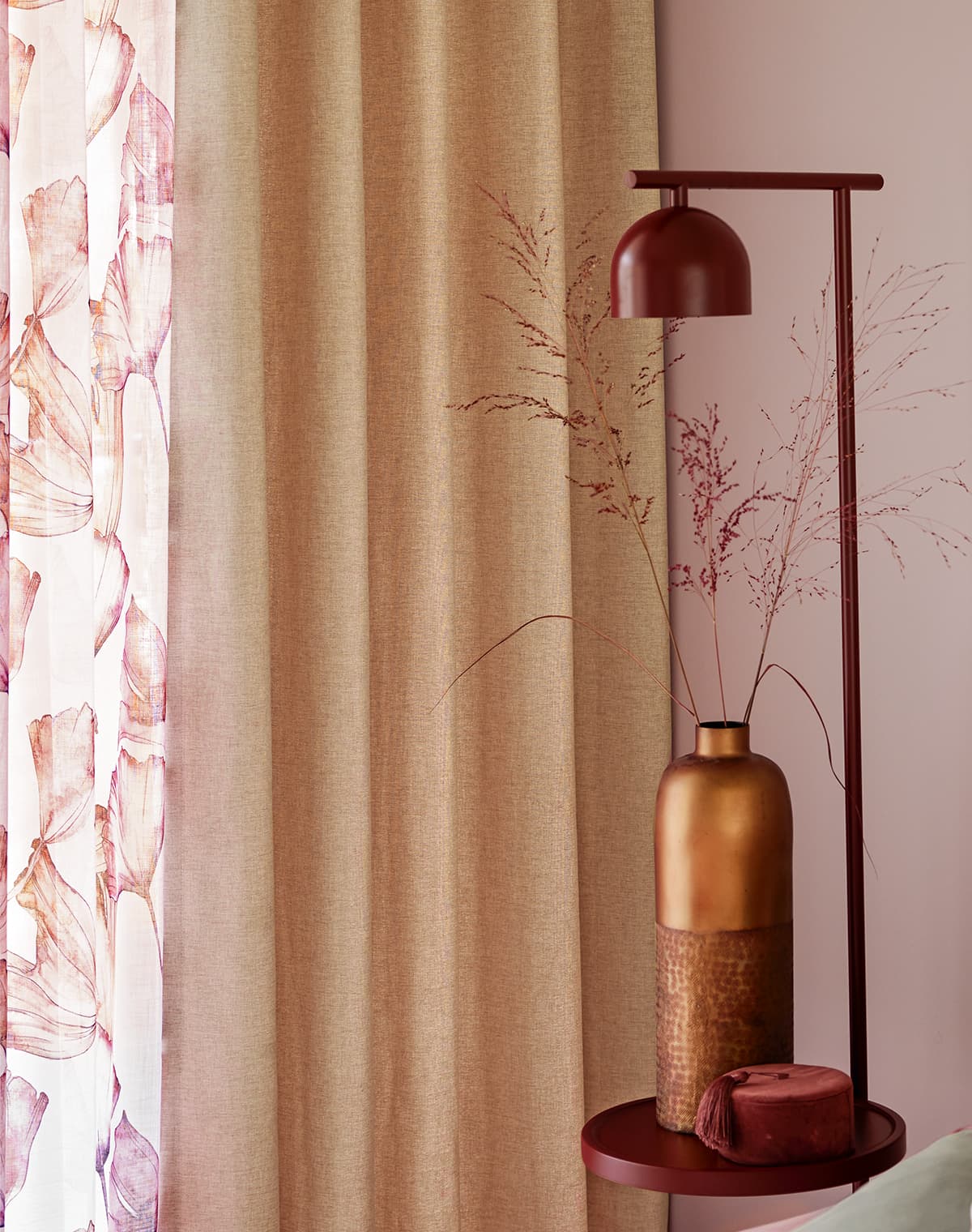 Das Bild zeigt Gardinen am Fenster in bronzefarbenem und rosa, floralem Design. In der Mitte steht ein dunkler roter Beistelltisch mit Dekorationsartikeln, ergänzt von einer roten Lampe, die die harmonische Farbgestaltung unterstreicht.