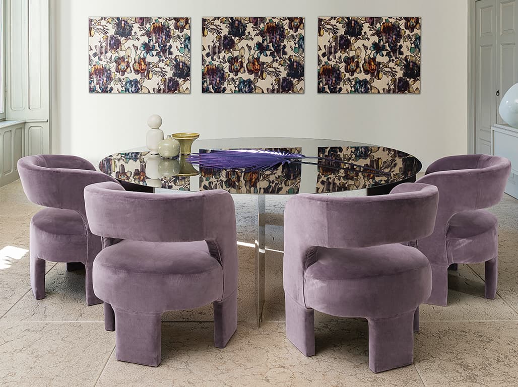 Das Bild zeigt einen spiegelnden Rundtisch, um den violette Stühle gruppiert sind. Der Tisch ist stilvoll dekoriert. Der Raum wirkt großzügig und luftig. An der Wand hängen Gemälde, die zur Atmosphäre passen und sich im Tisch spiegeln.