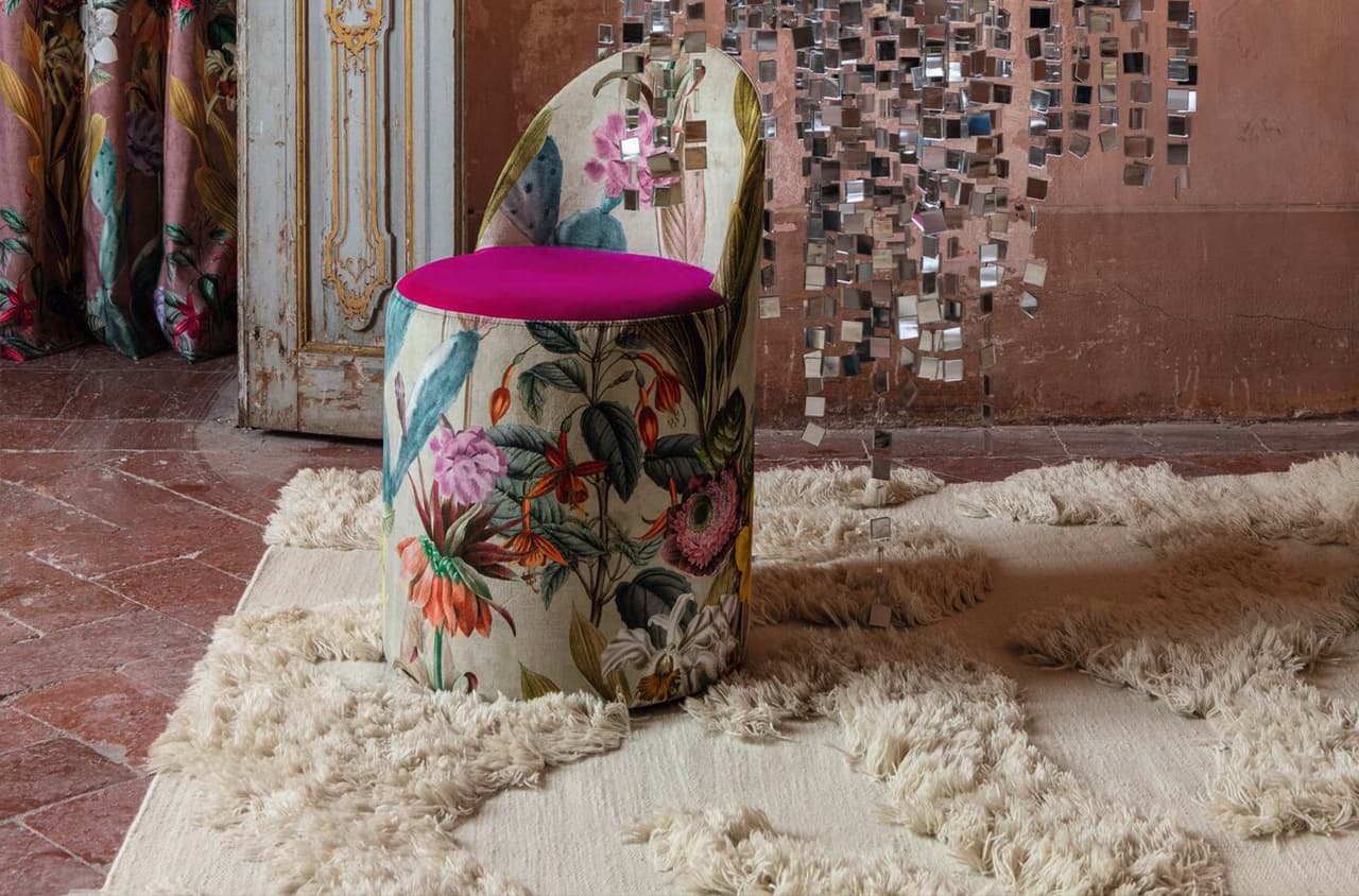 Ein Stuhl mit einem farbenfrohen, blumigen Muster und einer pinken Sitzfläche steht auf einem weißen, flauschigen Teppich in einem rustikalen Raum mit hängenden Spiegelmosaiken.