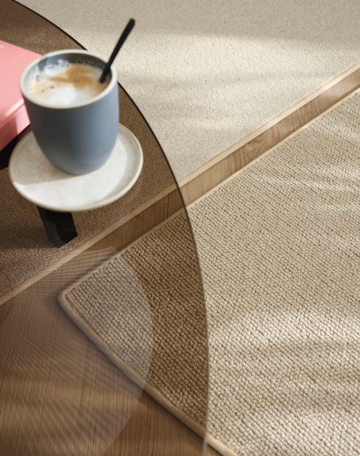 Eine Nahaufnahme von zwei ovalen Teppichen, die aneinander grenzen. Beide sind in beige gehalten und im Vordergrund ist ein Tisch mit einem Kafeebecher darauf.