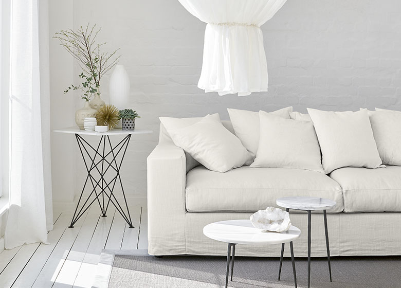 Das Bild zeigt ein weißes Sofa in einem hellen, weiß gehaltenen Raum. Ein Tisch mit Pflanzen sorgt für farbliche Abwechslung. Die Gesamtwirkung ist hell, harmonisch und minimalistisch.