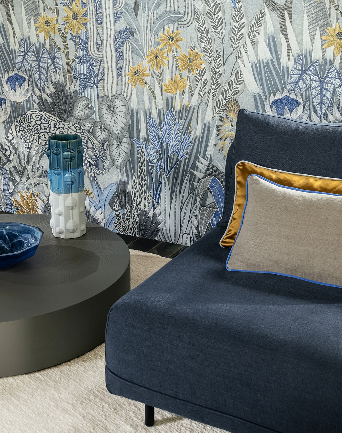 Blaues Sofa mit graum und goldenem Kissen vor einer Tapete mit floralem Printmotiv
