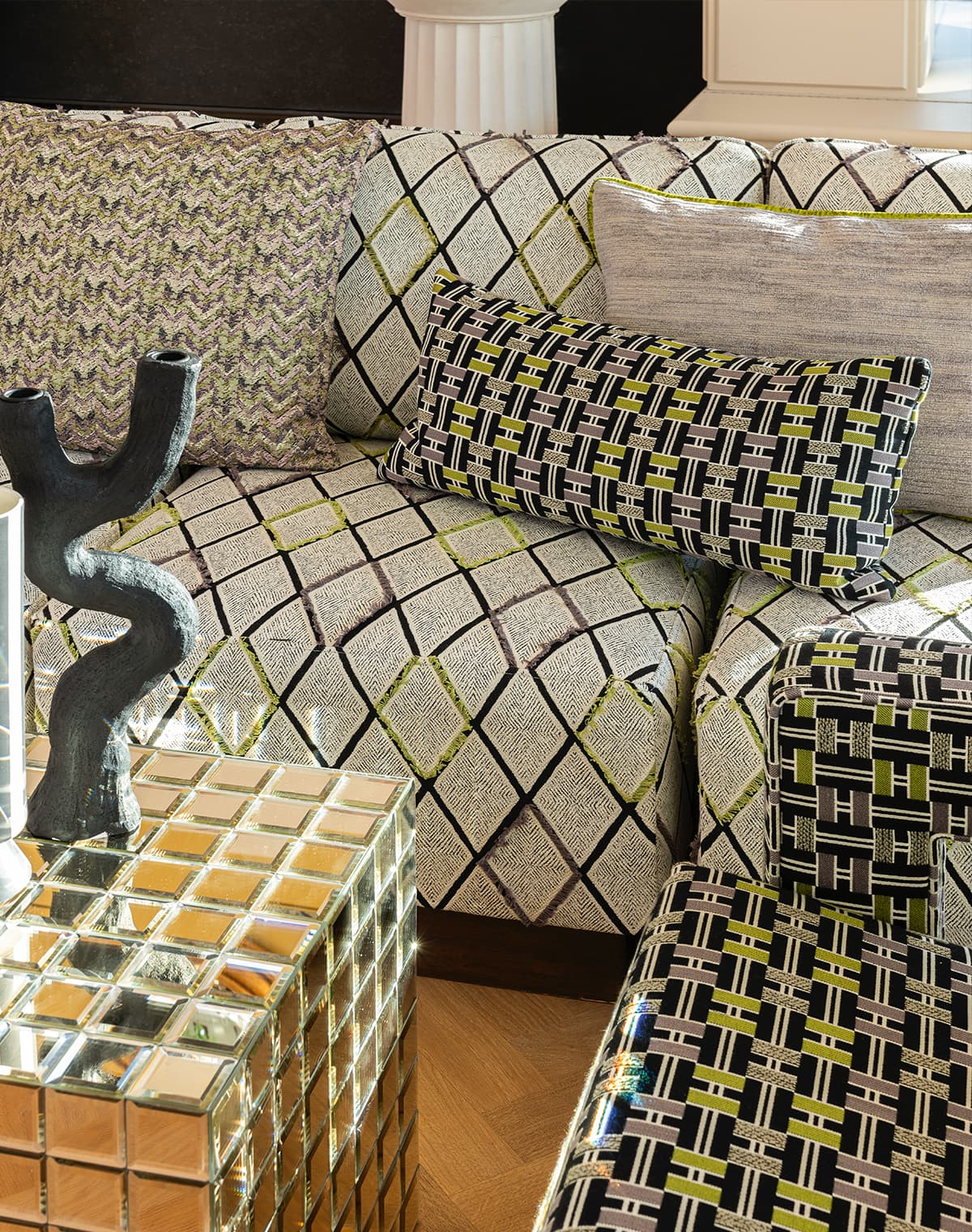 Das Bild zeigt grau-schwarz gemusterte Sofas mit grünen und lilafarbenen Akzenten im Wohnzimmer, ergänzt durch einen spiegelnden Würfeltisch. Die Farben harmonieren gut und schaffen eine angenehme Atmosphäre. 